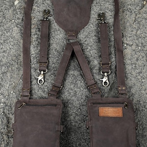 May include: A dark brown leather harness featuring two attached pouches with zippered closures. The system includes adjustable straps with metal swivel clips and a central back panel. A rectangular leather label with text is sewn onto the right pouch.
