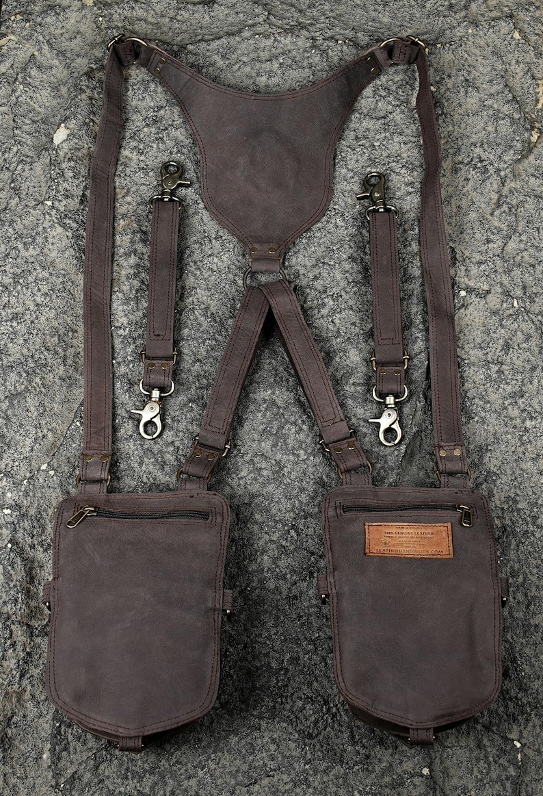 May include: A dark brown leather harness featuring two attached pouches with zippered closures. The system includes adjustable straps with metal swivel clips and a central back panel. A rectangular leather label with text is sewn onto the right pouch.