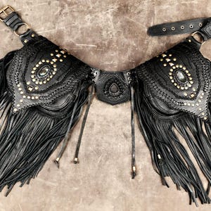 May include: A black leather belt featuring two large pouches with long fringe detailing. The pouches are adorned with metallic studs and a central oval gemstone. The belt includes an adjustable strap with a buckle closure and a central decorative piece.