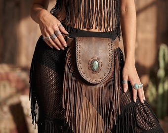 May include: A brown leather belt bag with a turquoise stone and fringe detailing. The bag is worn around the waist and has a flap closure. The bag is part of a bohemian style outfit.