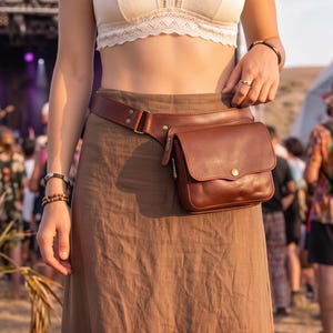 Op de afbeelding: Een donkerbruine leren heuptas met een rechthoekig hoofdvak, een voorflap met een metalen drukknoopsluiting en een verstelbare leren riem die rond de taille wordt gedragen over een lange bruine rok en een wit kort topje.