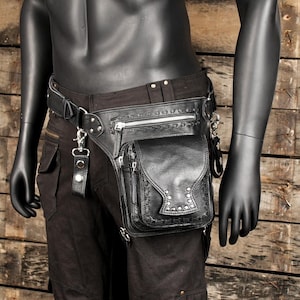 Puede incluir: Cinturón de cuero negro con una gran bolsa de cuero negro adjunta. La bolsa tiene cierre de cremallera y varios bolsillos. El cinturón tiene detalles plateados y una hebilla.