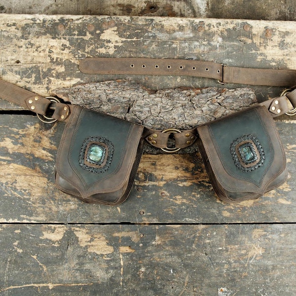 May include: A brown leather utility belt featuring two attached pouches with dark green leather accents. Each pouch is centered with a rectangular iridescent gemstone surrounded by decorative stitching. The belt includes metal rings and a buckle.