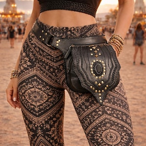 Puede incluir: Riñonera de cuero negro con tachuelas doradas y una piedra ovalada decorativa. El cinturón se lleva con pantalones estampados en tonos marrones y negros. La riñonera es un accesorio elegante.