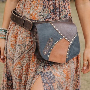 Puede incluir: Bolso cinturón de cuero marrón con diseño patchwork. El bolso presenta secciones de cuero marrón, beige y azul grisáceo, adornadas con pequeños tachuelas plateadas. El cinturón es de color marrón oscuro y tiene un cierre de hebilla. El bolso se lleva en la cintura.