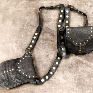 Op de afbeelding: Een zwarte holster-tas met slangenleer-textuur, bestaande uit twee tasjes verbonden door verstelbare riemen. De riemen zijn versierd met messingkleurige klinknagels en ronde hardware. Elk tasje heeft een klepsluiting met bijpassende klinknagels.
