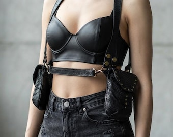 May include: A black leather-like bustier top paired with a black harness featuring two textured pouches on the sides. The harness includes metallic hardware, studs, and adjustable straps that connect the pouches to a central horizontal chest strap.