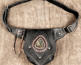 Puede incluir: Un bolso de cadera de cuero negro con correa ajustable y hebilla de latón, detalle lateral de cordones con dos tiras de cuero colgantes y una piedra iridiscente en forma de lágrima engastada en un borde trenzado de color cobre.