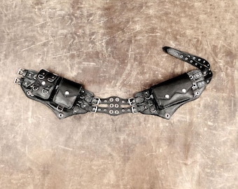 May include: A black utility belt featuring multiple attached pouches with silver-toned metal hardware, including buckles, rivets, and D-rings. The belt has a structured design with adjustable straps and reinforced eyelets along the central section.