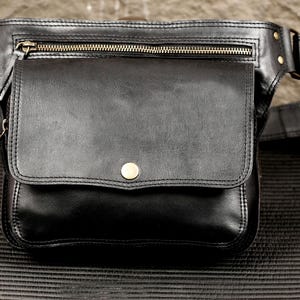 May include: A black leather waist bag featuring a large front flap with a single brass-toned snap closure, a horizontal zippered pocket above the main compartment, and an adjustable strap with brass-toned metal hardware and rivets.