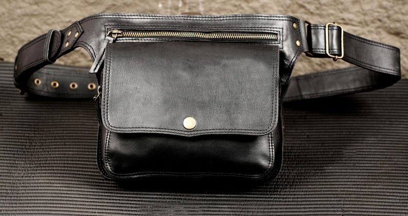 May include: A black leather waist bag featuring a large front flap with a single brass-toned snap closure, a horizontal zippered pocket above the main compartment, and an adjustable strap with brass-toned metal hardware and rivets.