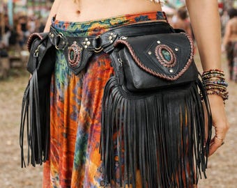 May include: Black leather belt bag with fringe and decorative stitching. The bag features two pouches and a central belt with metal hardware. The belt bag is worn over a colorful tie-dye skirt, creating a bohemian style.