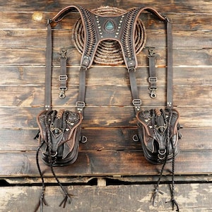 以下が含まれることがあります： 2つのポーチが付いた茶色の革製ハーネス。ハーネスは、複雑なステッチ、金属のアクセント、中央のターコイズストーンが特徴です。ポーチには編み込みのディテールと長い垂れ下がったコードが付いています。木製の背景に表示されています。