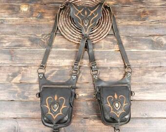 May include: A black leather dual holster bag featuring two symmetrical pouches with tan decorative scrollwork and central stone accents. The pouches are connected by adjustable straps to a central back piece with a matching tan design.