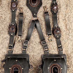 May include: A black leather harness featuring two attached pouches, adjustable straps with brass-toned hardware, and decorative dark oval stones framed by braided brown leather accents. The design includes multiple metal rings and clips.