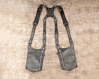 May include: A black leather shoulder holster harness featuring two vertical zippered pockets, adjustable straps with silver-toned metal grommets, and a central back panel. The design includes reinforced stitching and metal hardware connecting the straps.