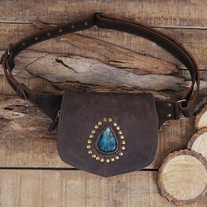 Puede incluir: Una riñonera de cuero marrón oscuro con una correa ajustable con ojales metálicos y una hebilla. La solapa frontal está decorada con una piedra preciosa azul en forma de lágrima rodeada por un patrón circular de pequeños remaches de latón.