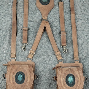 May include: A tan leather harness featuring two attached pouches and two detachable straps. Each pouch and the central back panel are adorned with an oval, iridescent blue-green stone set in a dark, scalloped leather frame with brass hardware.