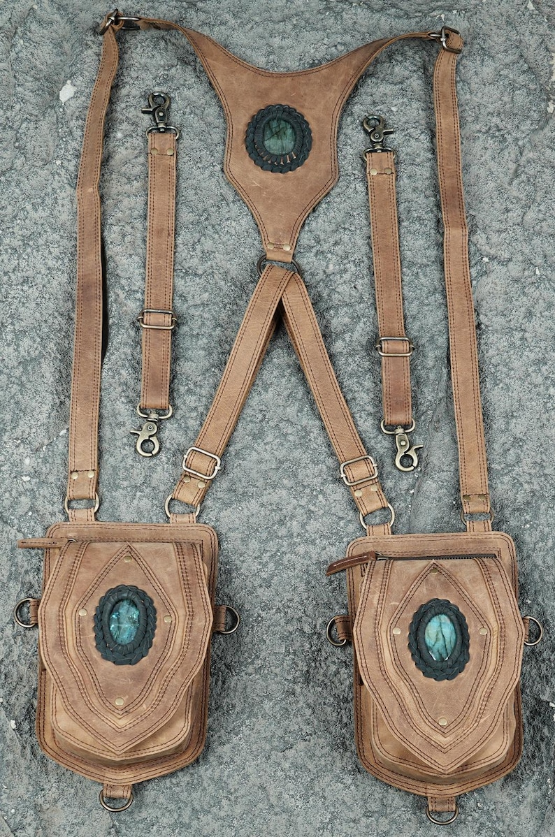 May include: A tan leather harness featuring two attached pouches and two detachable straps. Each pouch and the central back panel are adorned with an oval, iridescent blue-green stone set in a dark, scalloped leather frame with brass hardware.