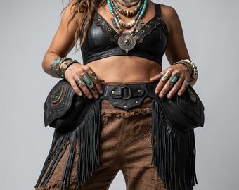 May include: A black leather belt featuring two large side pouches with long fringe detailing and a central metal ring. The belt is worn over brown trousers, paired with a black leather bralette, layered turquoise and beaded necklaces, and multiple silver rings.