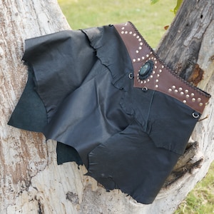 Peut inclure: Short en cuir noir avec une ceinture en cuir marron ornée d'une boucle en métal décorative et de clous. La ceinture a une pierre vert foncé au centre.