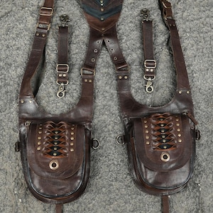 May include: A dark brown leather shoulder harness featuring two large pouches with woven lace-up detailing, brass-toned metal studs, and adjustable straps. The central back panel includes a dark green stone accent and metal hardware for secure attachment.