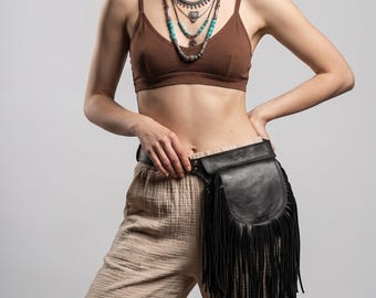 Könnte beinhalten: Eine schwarze Leder-Bauchtasche mit langen Fransen. Die Tasche wird mit einem schwarzen Gürtel um die Taille getragen. Das Bild zeigt auch einen braunen BH, mehrere Halsketten und eine beige Hose.