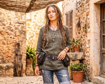 May include: An olive green long-sleeved linen shirt paired with blue denim jeans and a dark brown leather utility belt featuring two side pouches and a central stone embellishment, worn with a layered beaded necklace and multiple bracelets.