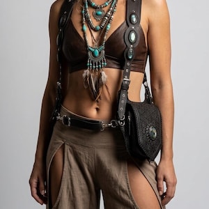 May include: A dark brown leather bralette paired with tan trousers featuring large thigh cutouts. A dark leather harness with silver studs and oval stones connects to a small black embossed leather pouch. Layered turquoise and silver necklaces.
