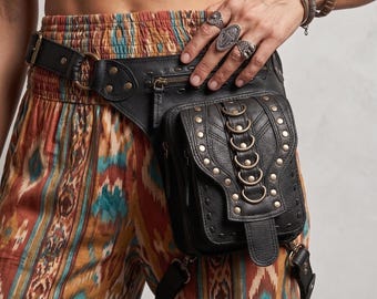 May include: A black leather utility belt bag featuring a main pouch with a flap closure, multiple brass-toned metal rings, and decorative studs. The bag includes an adjustable waist strap and a secondary leg strap, both secured with metal hardware.