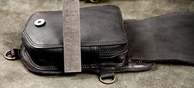 Metal ruler measuring side depth and zippered pocket thickness of black leather shoulder holster bag.