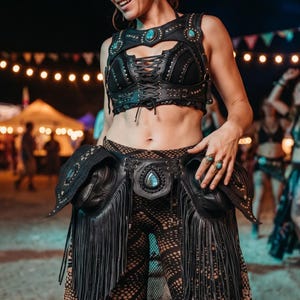 Könnte beinhalten: Schwarzes Leder-Festival-Outfit mit türkisfarbenen Akzenten. Das Outfit umfasst ein Schnür-Crop-Top, einen Hüftgürtel mit Fransen und eine gemusterte Hose. Das Bild wurde bei einer Outdoor-Veranstaltung mit Lichterketten aufgenommen.