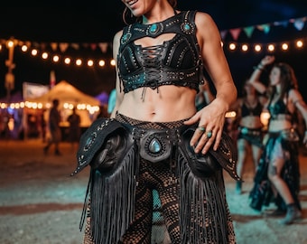 May include: Black leather festival outfit with turquoise accents. The outfit includes a lace-up crop top, a fringed hip belt, and patterned pants. The image is taken at an outdoor event with string lights in the background.