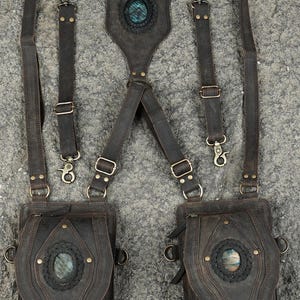 May include: A dark brown leather harness featuring two attached pouches, adjustable straps with metal clips, and three oval-shaped iridescent stones set into braided leather accents on the back panel and the front of each pouch.