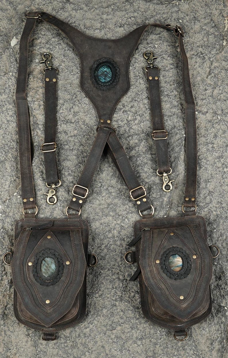 May include: A dark brown leather harness featuring two attached pouches, adjustable straps with metal clips, and three oval-shaped iridescent stones set into braided leather accents on the back panel and the front of each pouch.