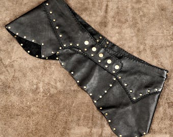 Puede incluir: Un cinturón envolvente de cuero negro con un diseño asimétrico, adornado con múltiples tachuelas metálicas doradas dispuestas en filas y a lo largo de los bordes. El cinturón incluye un panel ancho con un borde cosido reforzado.