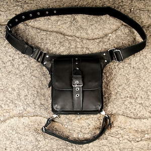 Op de afbeelding: Een zwarte leren been-tas met een rechthoekig hoofdvak, een klep aan de voorzijde, een verticale riem met metalen gesp en zilverkleurige hardware. De tas is voorzien van een verstelbare heupriem en een extra band voor rond het bovenbeen.