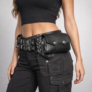 Könnte beinhalten: Ein schwarzer Utility-Gürtel mit einem breiten, lederartigen Band, doppelten silberfarbenen Schnallen, mehreren Metallösen und silbernen Ringen. Der Gürtel verfügt über eine rechteckige Tasche mit Klappverschluss und zwei Druckknöpfen.