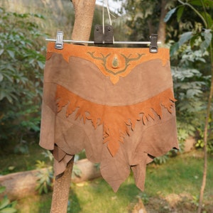 May include: A brown suede skirt featuring a layered design with darker brown and lighter tan panels. The waistband includes a decorative cutout detail resembling antlers surrounding a central teardrop-shaped accent. The hem has a jagged edge.