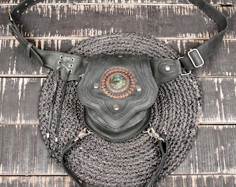 May include: A black leather belt bag featuring a central pouch with a circular brown stitched accent and a round iridescent stone inlay. The bag includes an adjustable strap with metal buckles, side lacing details, and two lower attachment clips.