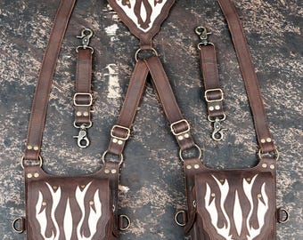 May include: A brown leather harness featuring two attached pouches and two detachable straps. The harness and pouches are decorated with white flame-like leather overlays. It includes brass-toned metal hardware, adjustable buckles, and swivel clips.