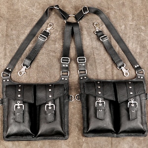 Könnte beinhalten: Ein schwarzes Leder-Doppeltaschen-Geschirr mit verstellbaren Riemen, silberfarbenen Metallschnallen, Nieten und Karabinerhaken. Die zwei rechteckigen Taschen haben jeweils einen Klappenverschluss mit Schnalle und sind durch einen Ring verbunden.