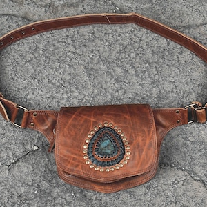 May include: A brown leather waist bag featuring an adjustable strap with a metal buckle. The front flap is decorated with a central teardrop-shaped blue stone, surrounded by a dark stitched border and a ring of small metallic studs.