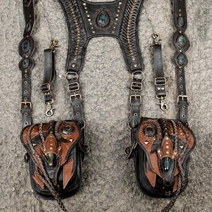 May include: A dark brown leather harness featuring two attached pouches, intricate braided detailing, and metallic studs. The harness includes adjustable straps with brass-toned buckles, metal clips, and embedded blue-toned stones on the shoulder straps.
