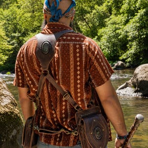 Peut inclure: Un harnais en cuir marron avec deux sacoches attachées, doté d'ornements métalliques ovales sur l'épaule et la sacoche latérale. Le harnais est porté sur une chemise à manches courtes rouille avec un motif géométrique blanc et un foulard bleu.