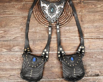 May include: A black leather harness featuring two symmetrical pouches with blue oval stones and brass studs. The upper triangular chest piece displays a white patterned inlay with a central blue stone, connected by adjustable straps and metal hardware.