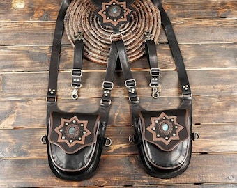 Puede incluir: Un arnés de cuero negro con dos bolsas de solapa, cada una adornada con una superposición de cuero marrón en forma de estrella y una piedra azul central. El arnés incluye correas ajustables, hebillas de metal y clips, con una pieza de cuero en forma de estrella en la parte superior central.