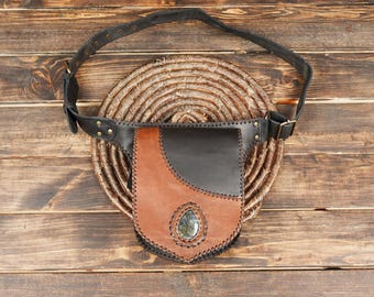May include: A leather hip bag featuring a two-tone design with a black upper section and a brown lower flap. The bag includes an adjustable black strap with metal hardware and a single teardrop-shaped iridescent stone centered on the front flap.