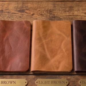 May include: A collection of leather journals in various colors: black, brown, light brown, dark brown, and white. Each journal is open to display the interior pages. The journals are displayed on a wooden surface.