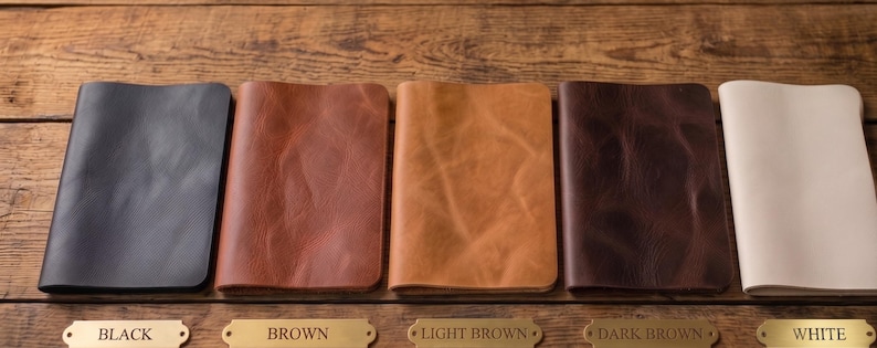 May include: A collection of leather journals in various colors: black, brown, light brown, dark brown, and white. Each journal is open to display the interior pages. The journals are displayed on a wooden surface.